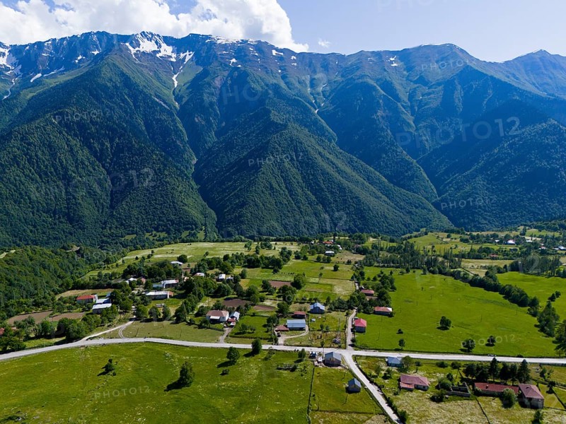 A small village in the middle of a vast green landscape with high mountains in the background, aerial view, landscape near Iskari, Mingrelia and Upper Svaneti, Samegrelo-Zemo Svaneti, Svaneti, High or Greater Caucasus, Georgia