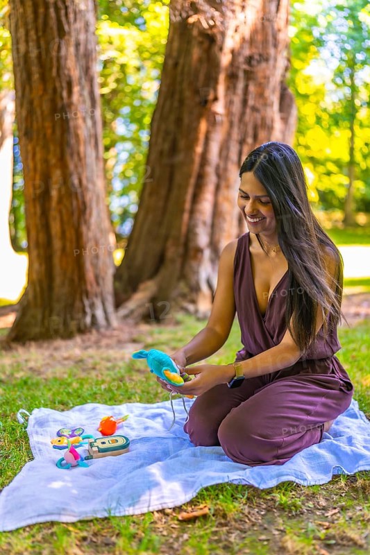 Happy young latin mother kneeling on a blanket in a park, preparing vibrant baby toys while enjoying the sunny outdoor atmosphere