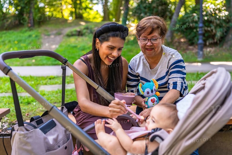 Grandmother, mother and baby girl enjoying time together at the park, feeding the baby girl and playing with a plush toy