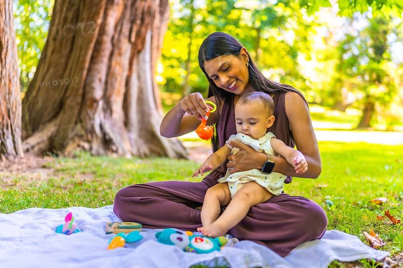 Latin mother holding her baby daughter and playing with colorful toys while sitting on a blanket in a park