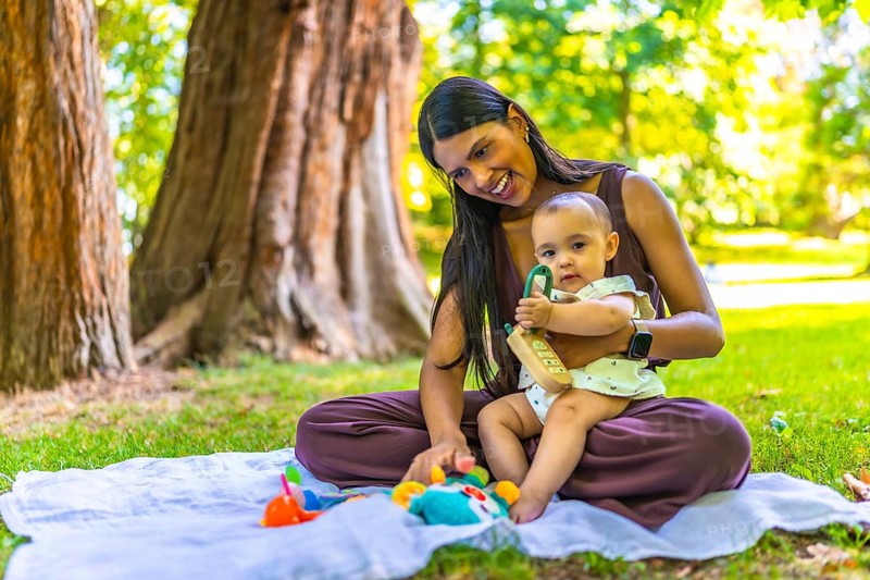 Latin mother and her baby daughter enjoying playtime with colorful toys on a blanket, under the shade of trees in a vibrant park