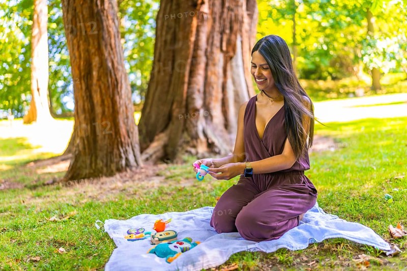 Smiling latin mother arranging baby toys on a blanket in a park, enjoying a sunny day outdoors