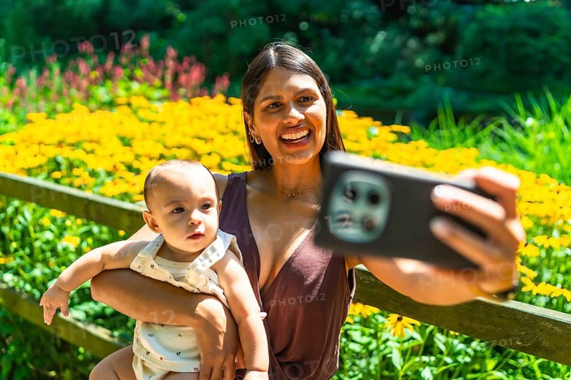 Happy latin mother holding her baby daughter and taking a selfie with smartphone in a beautiful garden full of yellow flowers