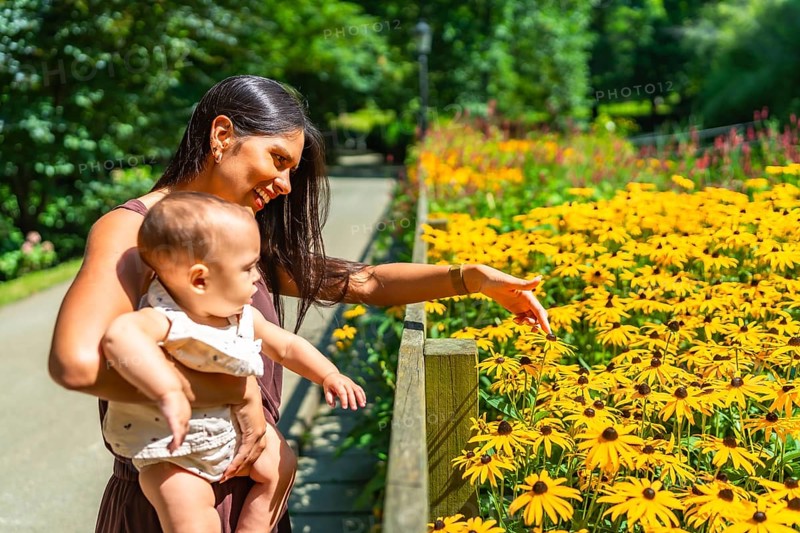 Latin mother holding her baby daughter and showing her yellow flowers in bloom in a sunny botanical garden