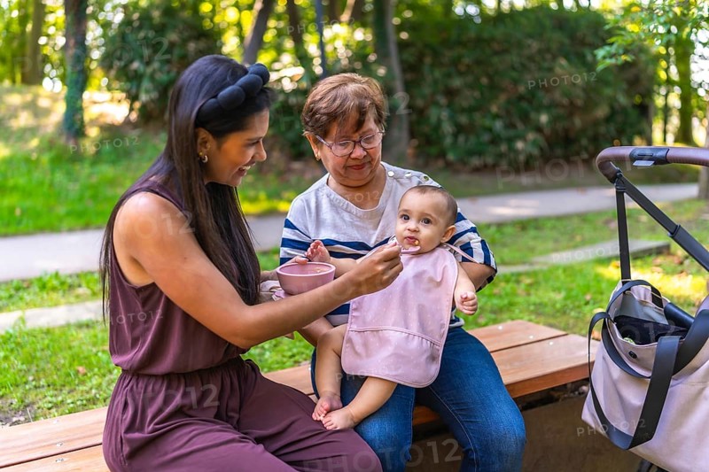 Mother feeding her baby girl solid food, with grandmother holding her, sitting on a park bench
