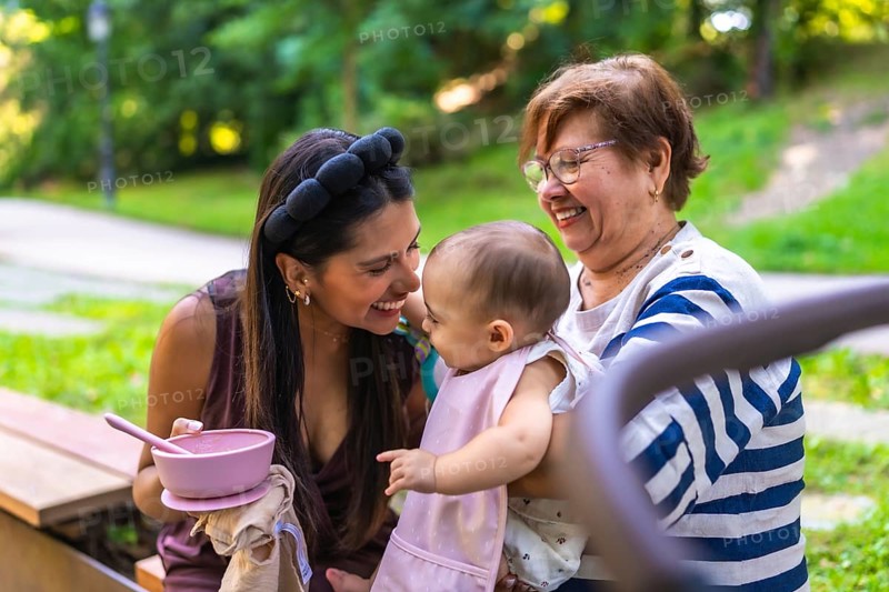 Grandmother holding her granddaughter while mother is feeding her in a park, enjoying family time together