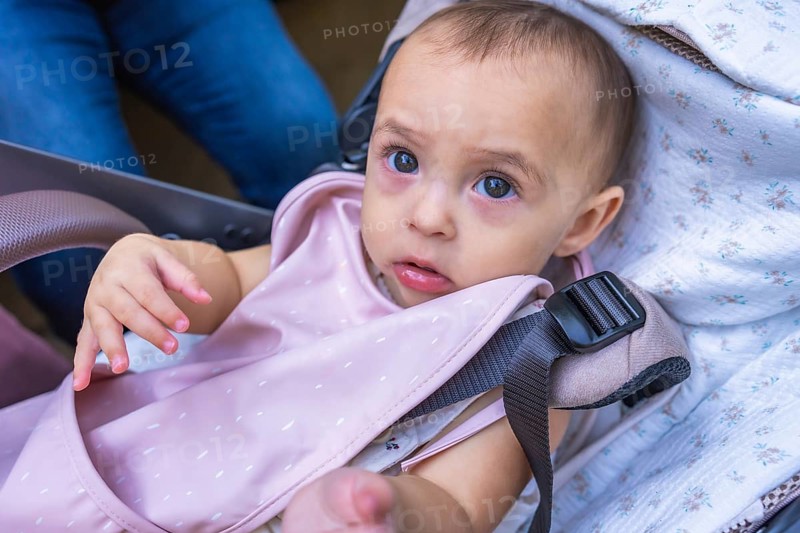 Adorable baby girl sitting in a stroller, secured with a safety harness and wearing a pink bib
