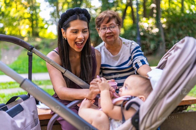 Grandmother, mother and baby girl enjoying a sunny day at the park, feeding the baby sitting in a stroller