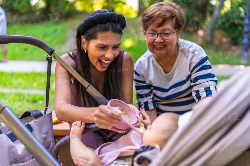 Latin mother feeding her baby girl solid food from a pink bowl, with grandmother assisting, in stroller at park