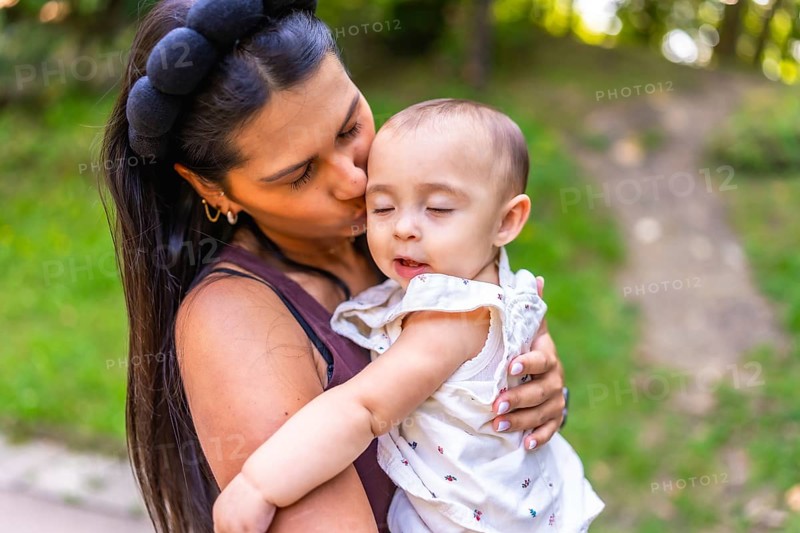 Young latin mother kissing her baby daughter while holding her lovingly in her arms, surrounded by the beauty of a green park