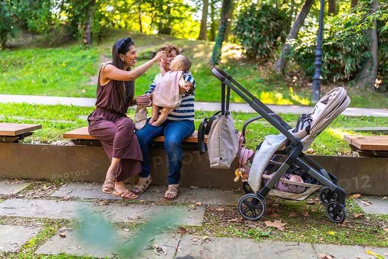 Three generations of a latin family enjoying time together in a park, with mother feeding her baby while sitting next to grandmother on a bench