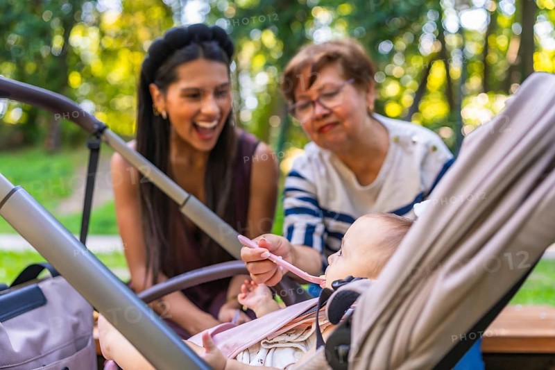 Grandmother feeds baby girl sitting in stroller, while mother watches, enjoying family time together in park