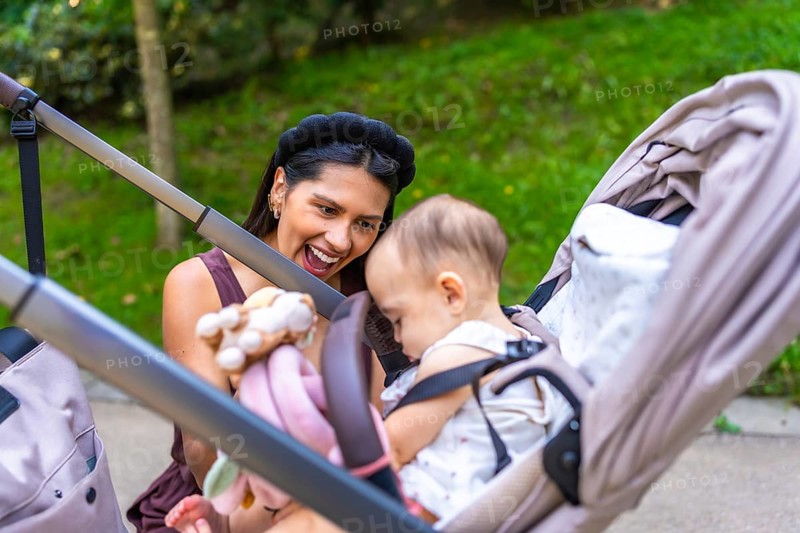 Happy latin mother showing toys to her baby daughter sitting in a stroller, enjoying a sunny day together in a park