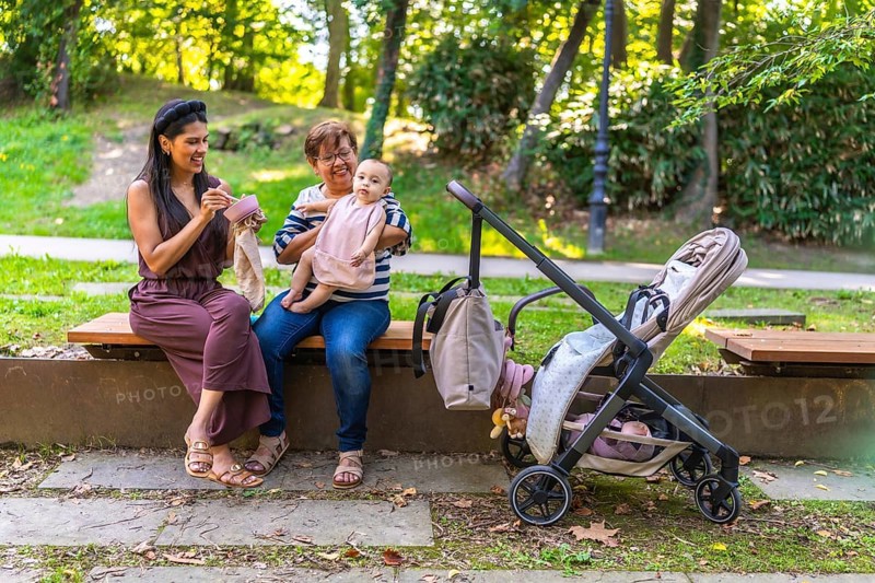 Grandmother holding baby girl while mother is eating and stroller is parked next to them on bench in park
