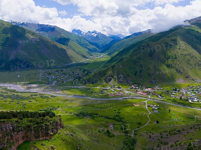 Green valley with river and village, surrounded by high mountains and cloudy sky, aerial view, view into the valley of the Snostskali River and the Terel River, Toti, Kazbegi, Qasbegi, Stepantsminda, Georgian Military Road, Mtskheta-Mtianeti Province, High Caucasus, Georgia