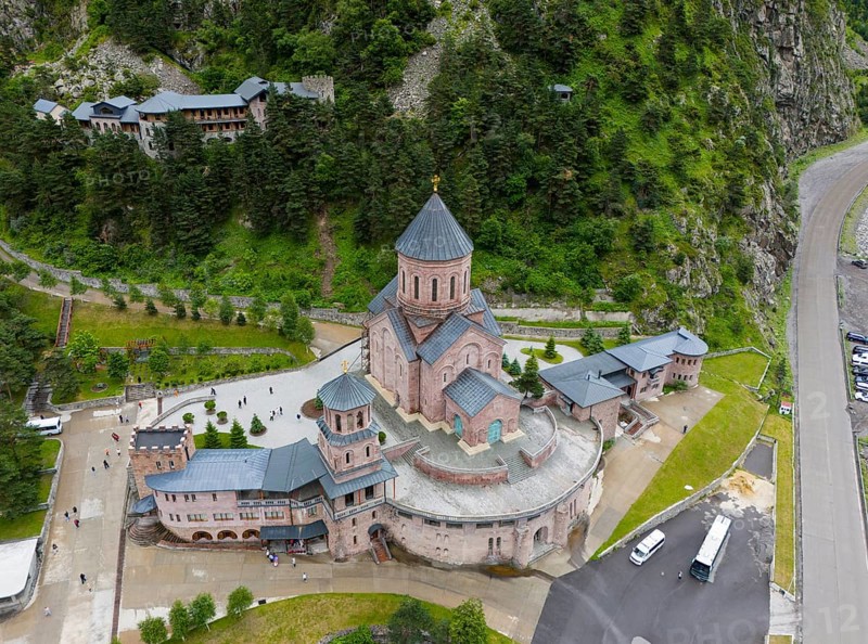 Large red brick church embedded in a wooded mountain landscape under a slightly cloudy sky, aerial view, Dariali Monastery complex, located in the Dariali Gorge of the Tergi River, Terek, Khevi Mountain Region, Georgian Military Road, Mtskheta-Mtianeti Province, High Caucasus, Georgia
