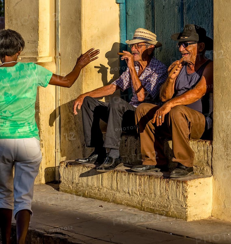 Two old Cubans smoke cigars and sit on a staircase in the warm light of the sunset, they greet a passer-by in a green T-shirt, Trinidad, Cuba, Sancti Spíritus, Sancti Spíritus