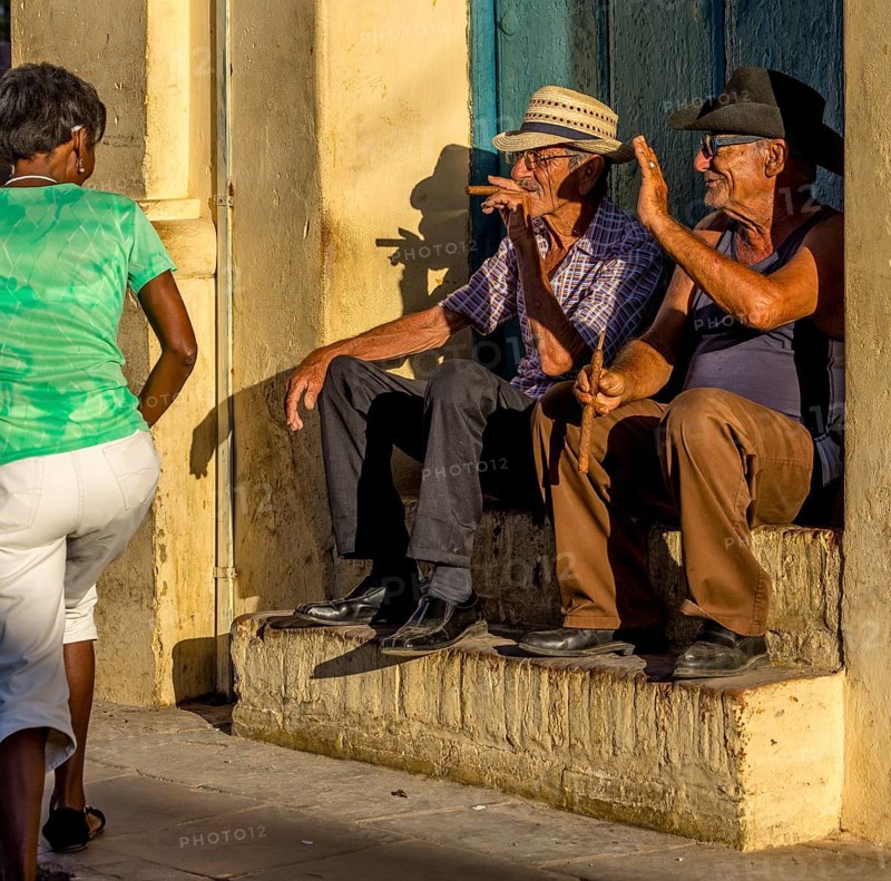 Two old Cubans smoke cigars and sit on a staircase in the warm light of the sunset, they greet a passer-by in a green T-shirt, Trinidad, Cuba, Sancti Spíritus, Sancti Spíritus