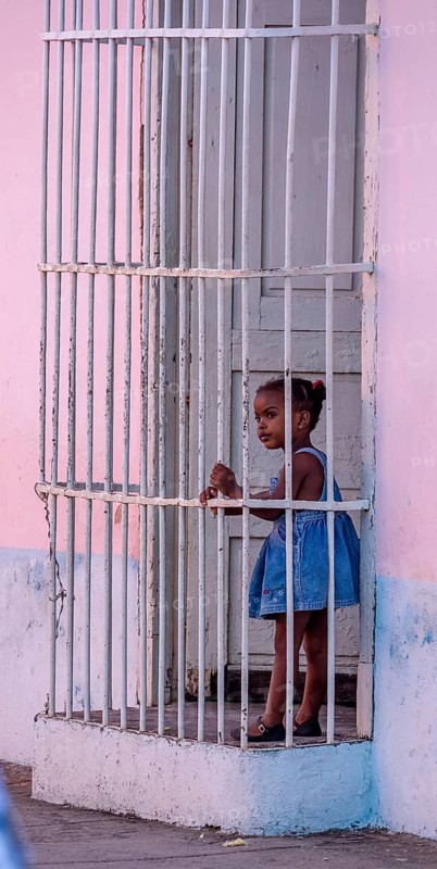 Little coloured girl behind window bars in a pink house, street of Trinidad, Cuba, Sancti Spíritus