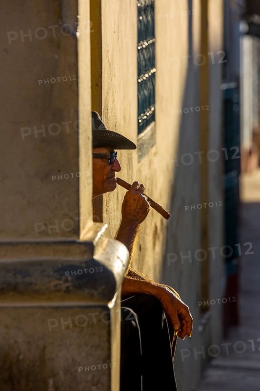 An old Cuban smokes a cigar and sits on a staircase in the warm light of the sunset, Trinidad, Cuba, Sancti Spíritus, Sancti Spíritus