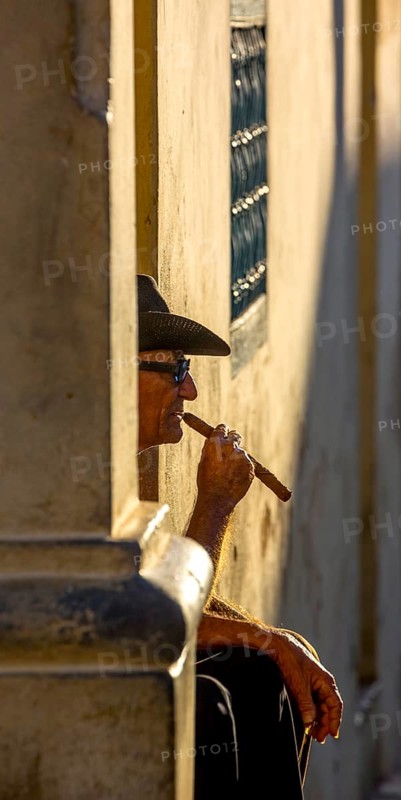 An old Cuban smokes a cigar and sits on a staircase in the warm light of the sunset, Trinidad, Cuba, Sancti Spíritus, Sancti Spíritus