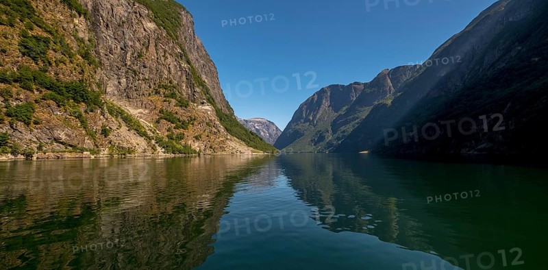 An approaching excursion boat on a beautiful fjord reflecting the light, surrounded by rocks partly overgrown with trees, above a bright blue sky. Gudvangen, Sogn og Fjordane, Norway, Scandinavia, Europe, Gudvangen, NOR, travel, tourism, destination, sightseeing, sea voyage, Hurtigruten, Hurtigruten trip, tourism, Norwegian, Scandinavian, tourist, cruise, adventure trip, mountains, tourist trip, boat trip, river, rocks, tourist ship, water, waters