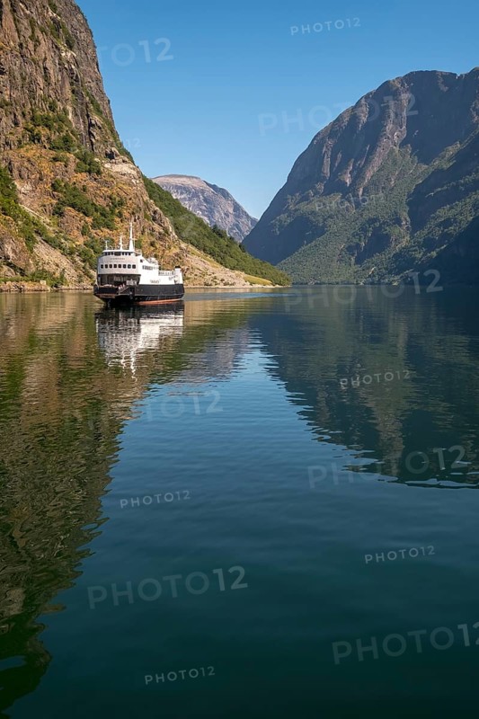 An approaching excursion boat on a beautiful fjord reflecting the light, surrounded by rocks partly overgrown with trees, above a bright blue sky. Gudvangen, Sogn og Fjordane, Norway, Scandinavia, Europe, Gudvangen, NOR, travel, tourism, destination, sightseeing, sea voyage, Hurtigruten, Hurtigruten trip, tourism, Norwegian, Scandinavian, tourist, cruise, adventure trip, mountains, tourist trip, boat trip, river, rocks, tourist ship, water, waters