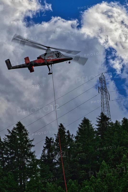 A helicopter flies over a forest and transports a load next to power lines, transport helicopter, timber transport, Saas Grund, Canton Valais, Switzerland