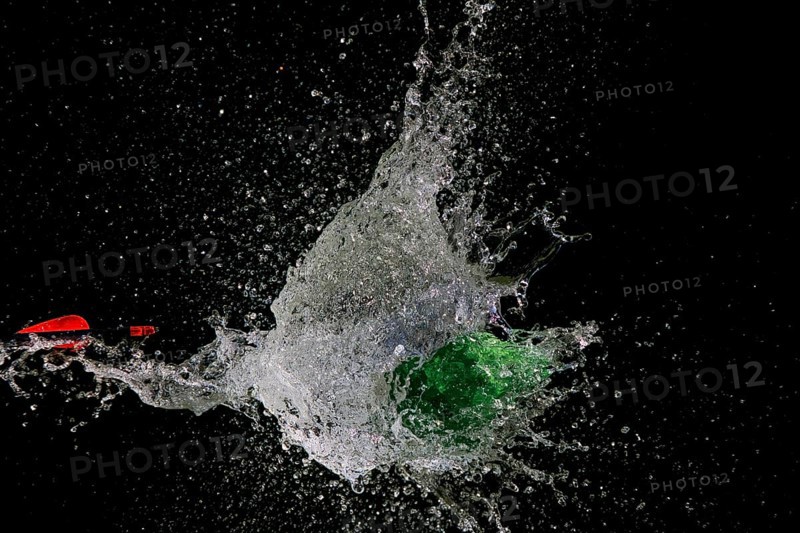 A green water balloon bursts after arrow penetration, causing water to splash, in front of a dark background, high-speed shot, water balloon, arrow