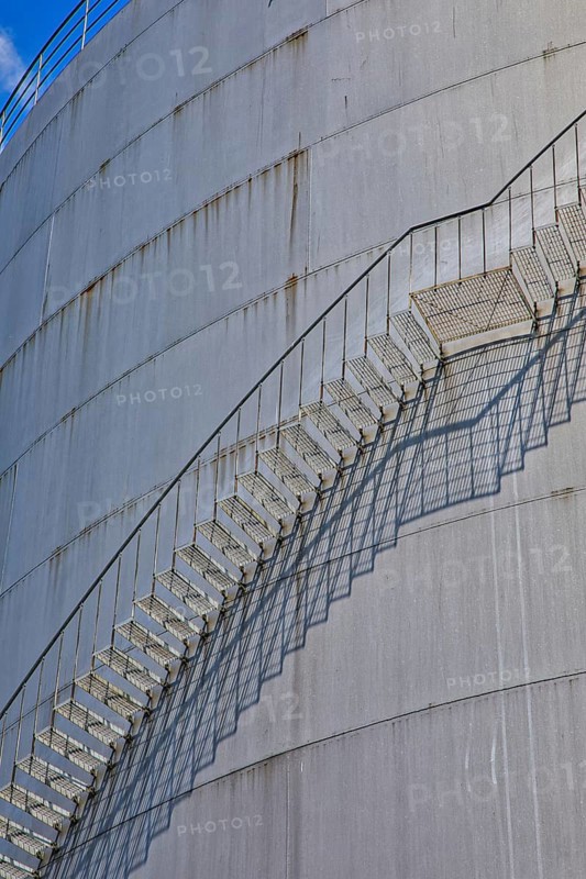 A long metal ladder stretches along a large cylinder building with shadows in the background, elevated tank, chemical, company, Grenzach- Wyhlen, Baden-Württemberg, Germany