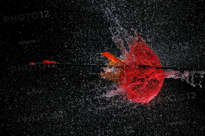 An arrow pierces a red water balloon and creates an impressive water explosion against a dark background, high-speed shot, water balloon, arrow