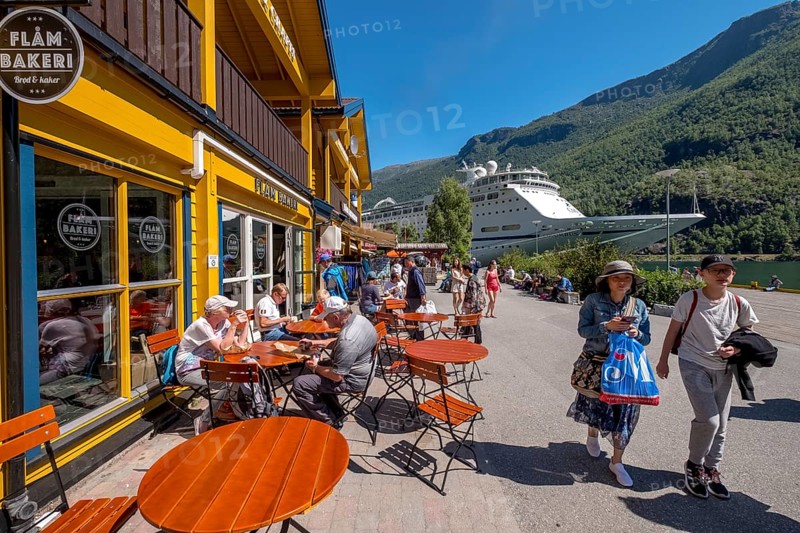 Tourists in Flåm, FABRIK KUTSALG FACTORY OUTLET, café, yellow wooden house, excursion ship, fjord, mountain, trees, blue sky, Sogn og Fjordane, Norway, Scandinavia, Europe, Flåm, NOR, travel, tourism, destination, sightseeing, sea voyage, Hurtigruten, Hurtigruten voyage, tourism, Norwegian, Scandinavian, tourist, cruise, adventure trip, pedestrian, river, forests, shipping, shops, shopping, shopping, signs, flags, tables, chairs, people, windows, shops, balconies