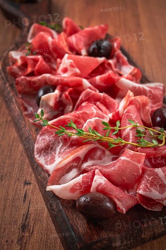 Freshly cut dried meat, served with olives and herbs, on a wooden chopping board, no people