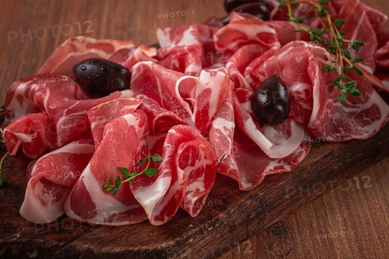 Freshly cut dried meat, served with olives and herbs, on a wooden chopping board, no people