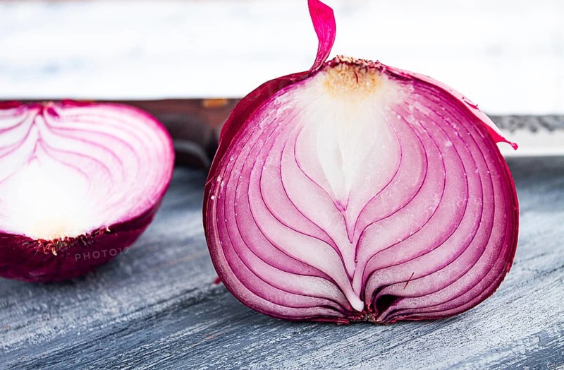 A vibrant red onion is cut in half, on a rustic wooden chopping board beside a sharp knife, ready for cooking