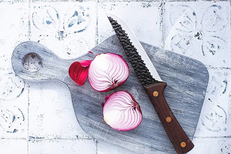 A vibrant red onion is cut in half, on a rustic wooden chopping board beside a sharp knife, ready for cooking