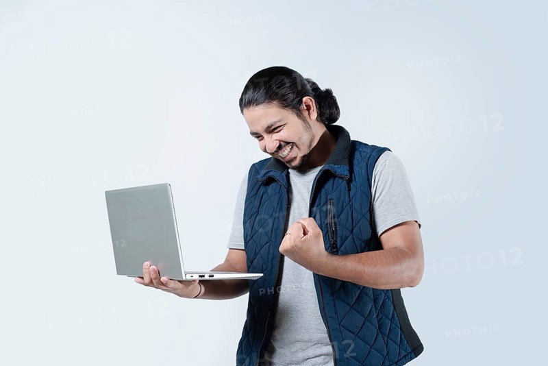 Male celebrating with fist in the air holding a laptop on white background. Young man celebrating holding a computer on isolated background