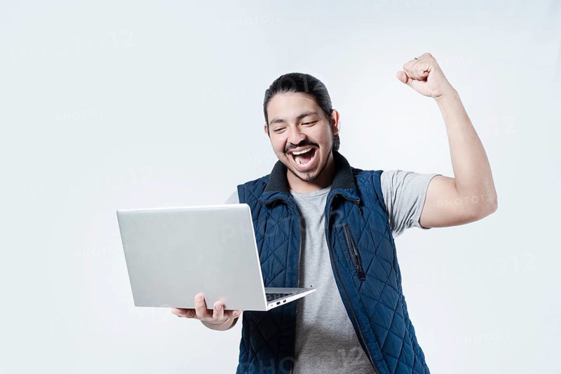 Young man celebrating holding a computer on isolated background. Male celebrating with fist in the air holding a computer on white background