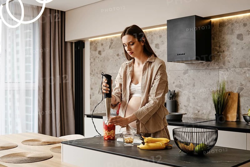 In a stylish kitchen, a pregnant woman blends fresh fruits like bananas and strawberries. Sunlight fills the space as she focuses on creating a nutritious smoothie, promoting a healthy lifestyle