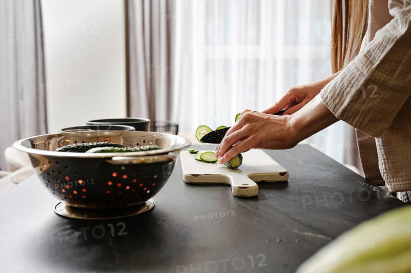 A person slices cucumbers on a cutting board in a well-lit modern kitchen. A colander rests nearby with fresh greens, emphasizing the focus on healthy cooking and meal preparation