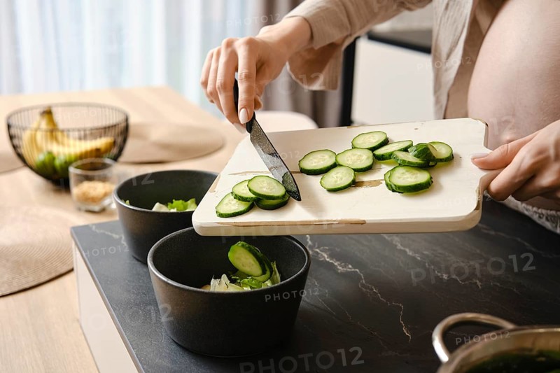 A person slices cucumbers on a wooden cutting board, adding them to two black bowls filled with salad ingredients