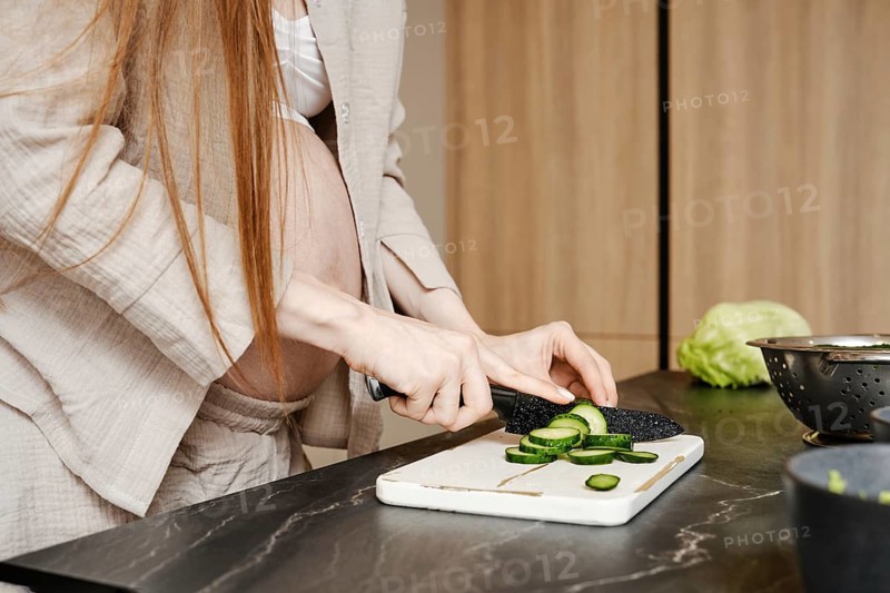 In a stylish kitchen, a pregnant woman with long hair cuts cucumbers on a white cutting board. She wears casual clothes and shows focus as she prepares healthy food, surrounded by fresh produce