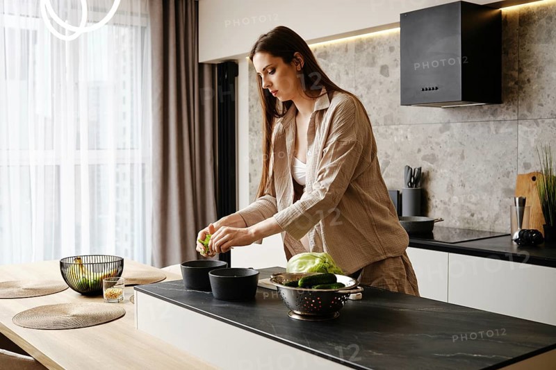 A woman with long hair is slicing vegetables in a contemporary kitchen. She focuses on her task while standing at a stone countertop