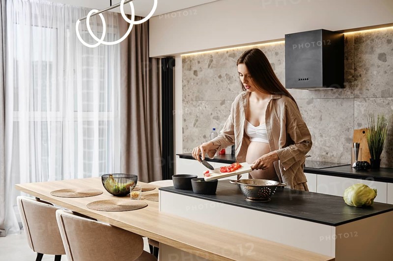 A pregnant woman with long hair is preparing a meal in a contemporary kitchen. She is cutting vegetables, showcasing healthy cooking. Natural light fills the room, enhancing a calm atmosphere