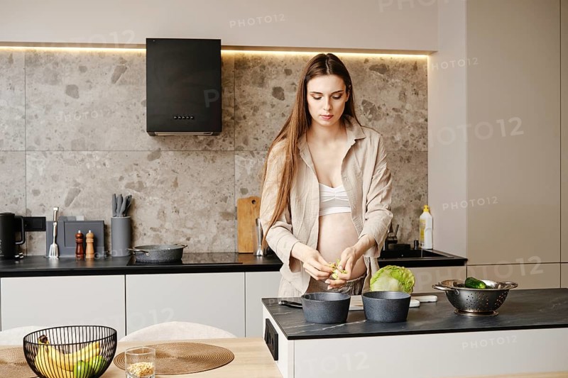 A pregnant woman stands in a stylish kitchen, chopping vegetables. She wears a casual outfit and showcases a serene demeanor. The sunlight enhances the contemporary decor and creates a warm ambiance