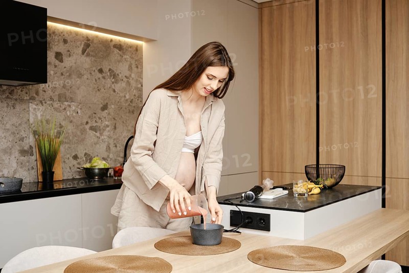 A woman in early pregnancy mixes ingredients in a pot while standing in her stylish kitchen. She pours fruit smoothie in a bowl