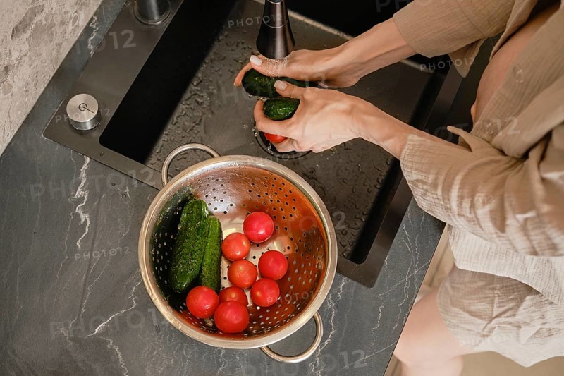 Hands scrub cucumbers under running water at a sleek kitchen sink. A colander nearby holds ripe tomatoes and cucumbers, showcasing a vibrant assortment of fresh produce