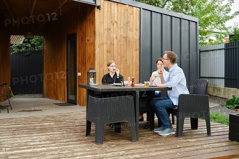 A family gather around a black table on a wooden deck of tiny house, sharing drinks and laughter in a stylish backyard. The surrounding greenery complements the contemporary design of the space