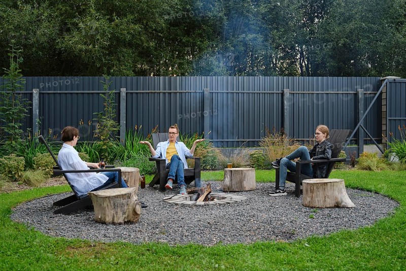 A family enjoy a relaxing evening around a fire pit in a backyard. They are sitting on wooden stools, engaged in conversation while the gentle smoke rises into the cool evening air