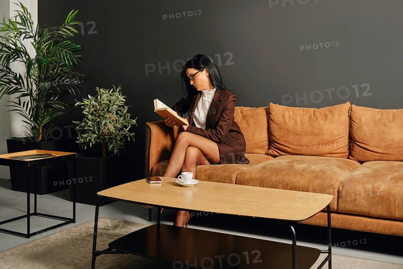 A woman dressed in a smart outfit is enjoying a quiet moment with a book on a cozy couch. The modern living room features greenery and minimalist furniture, creating a calm atmosphere